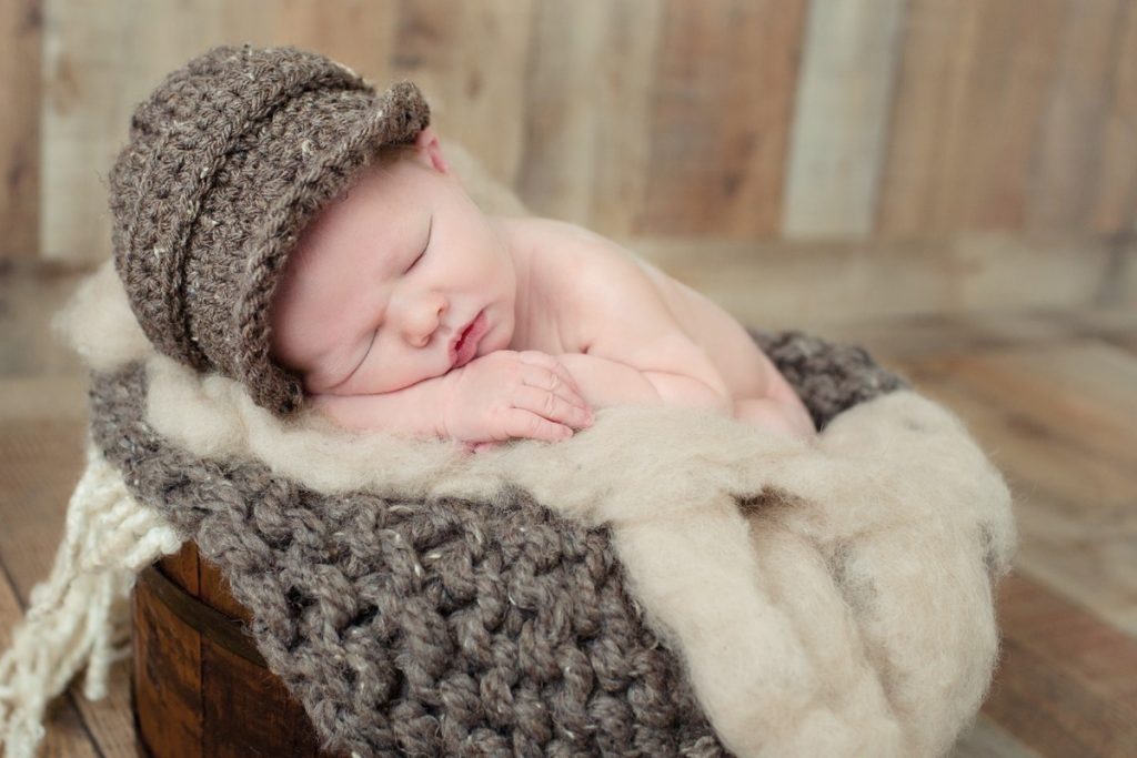 Newborn boy with brown knitted hat and chunky knit blanket in bucket of wool fluff