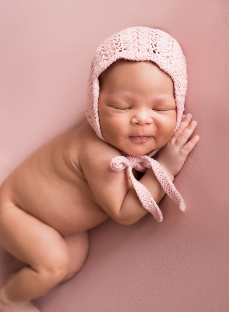 Babygirl grins posed onher side in Newborn Photosession on Soft Pink Baby Blanket