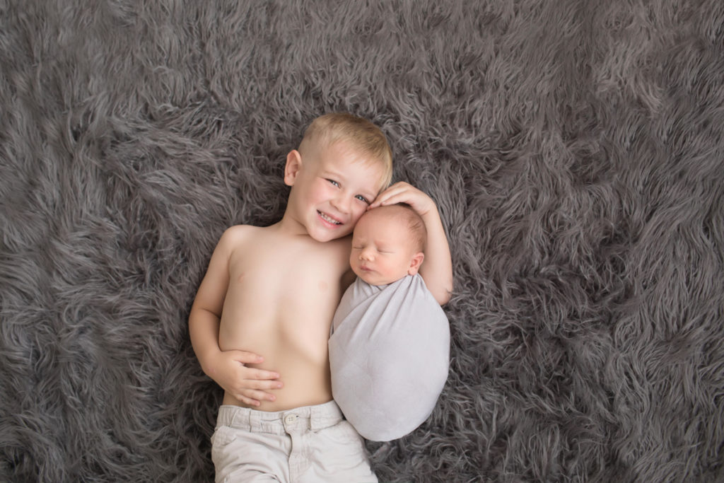 Big brother toddler cuddling newborn baby brother wrapped in grey posed on grey rug in Gainesville Florida