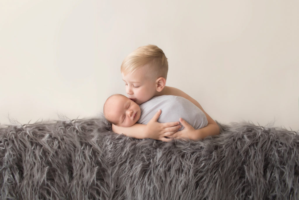 Toddler cuddling and kissing baby brother newborn wrapped in grey blanket on grey rug in Gainesville Florida