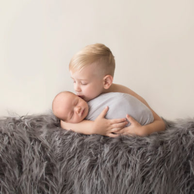 Toddler cuddling and kissing baby brother newborn wrapped in grey blanket on grey rug in Gainesville Florida