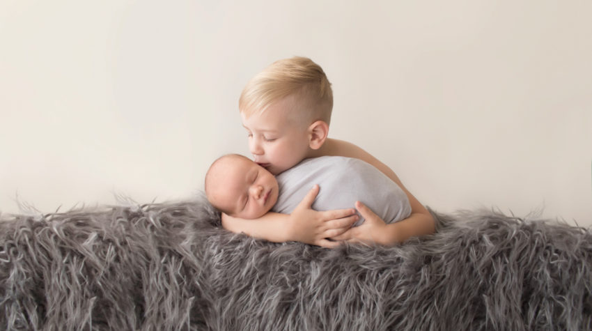 Toddler cuddling and kissing baby brother newborn wrapped in grey blanket on grey rug in Gainesville Florida