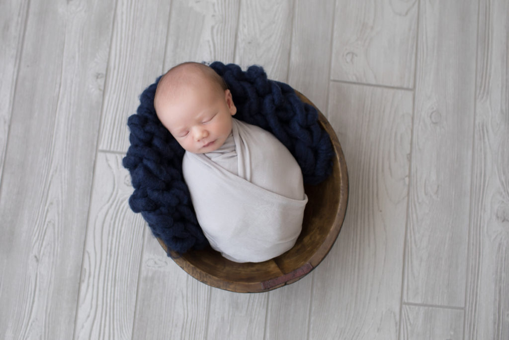 Baby newborn boy Chase wrapped in grey with navy knit blanket in brown bowl on grey wood