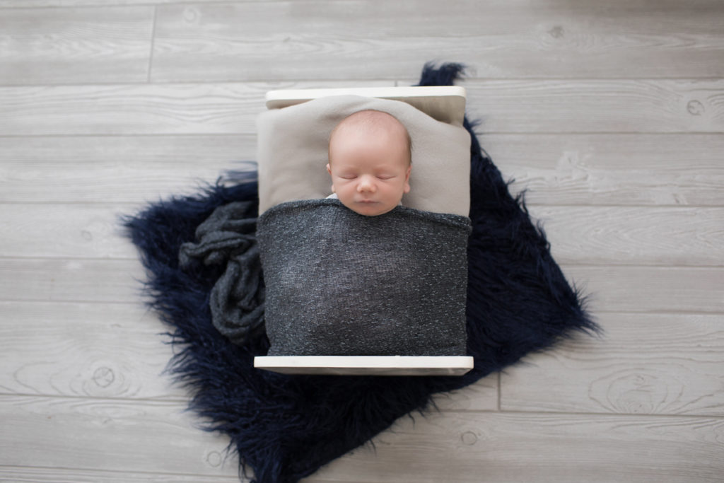 Baby newborn boy Chase tucked into white bed with grey blanket on navy rug in Gainesville Florida