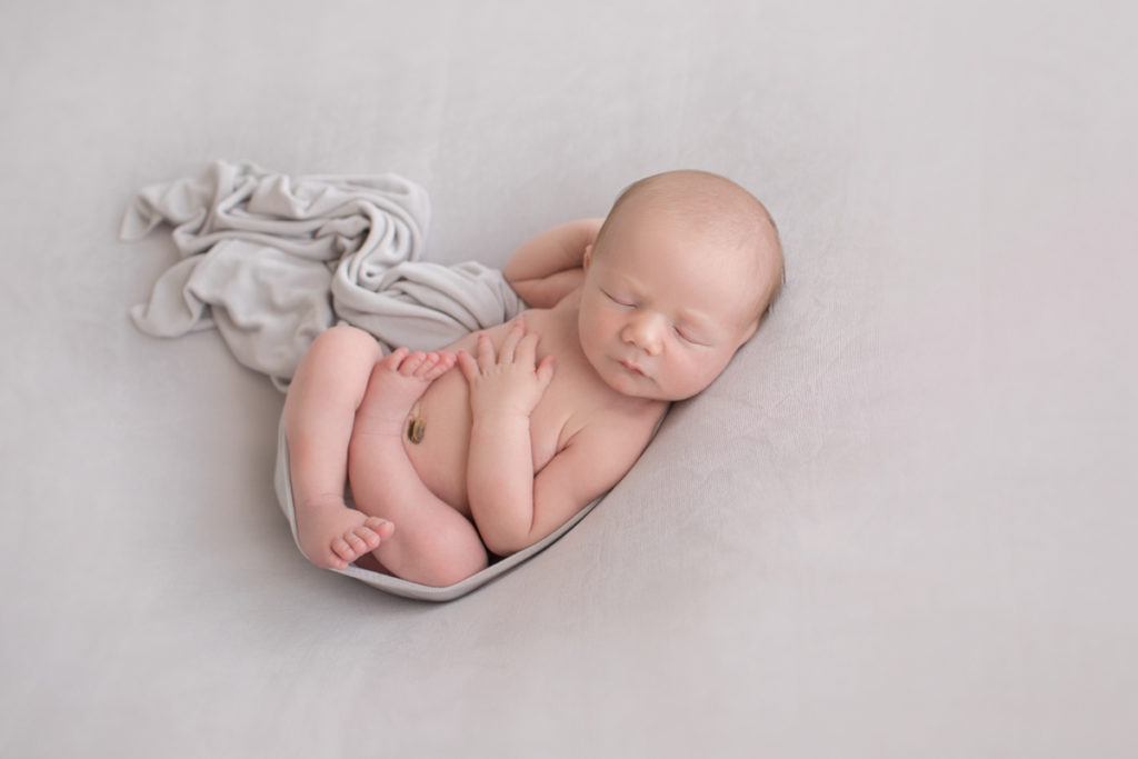 Naked baby newborn boy Chase with his arms and legs wrapped in front of him on grey blanket in Gainesville Florida