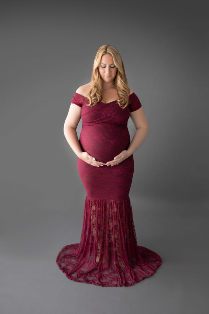 Pregnant mom in burgundy lace mermaid gown poses looking at her round belly anticipating having a baby girl soon