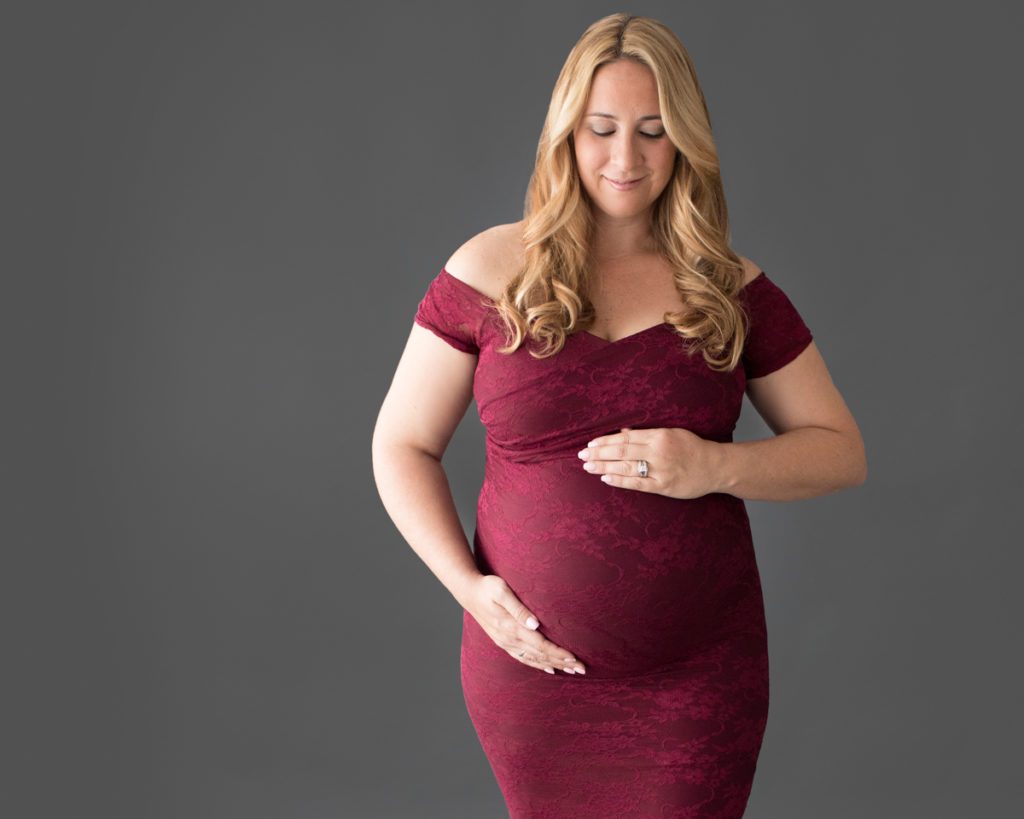 Pregnant mom in burgundy lace mermaid gown poses smiling at her round belly anticipating having a baby girl soon