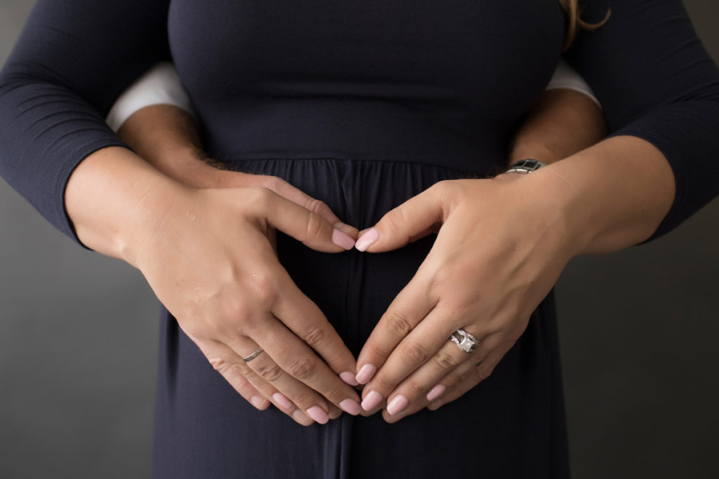 Moms hands on dads hands in the shape of a heart formed over pregnant belly navy maternity gown