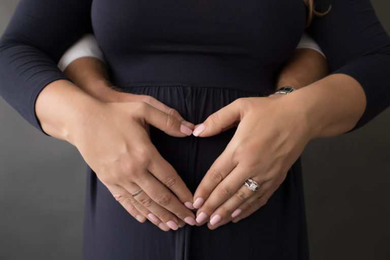 Moms hands on dads hands in the shape of a heart formed over pregnant belly navy maternity gown pictures Gainesville Florida