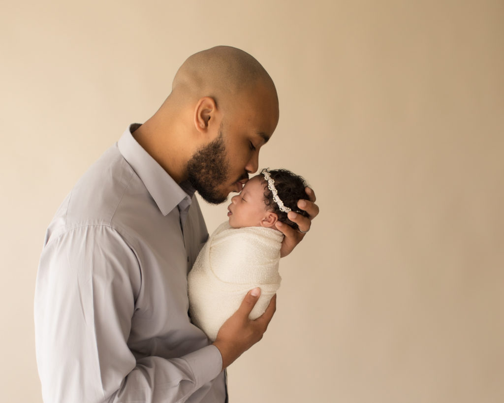 New Dad tenderly kissing newborn daughter Ayana wrapped in crea, and pearl headband
