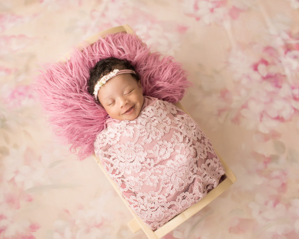 Newborn Girl Ayana with big smiles wrapped in pink lace shades on pink floral blanket in baby bed