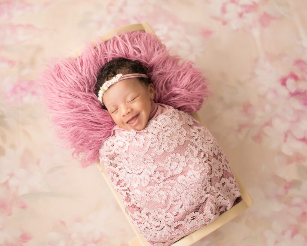 Newborn Girl Ayana with smiles and laughter wrapped in pink lace shades on pink floral blanket in baby bed