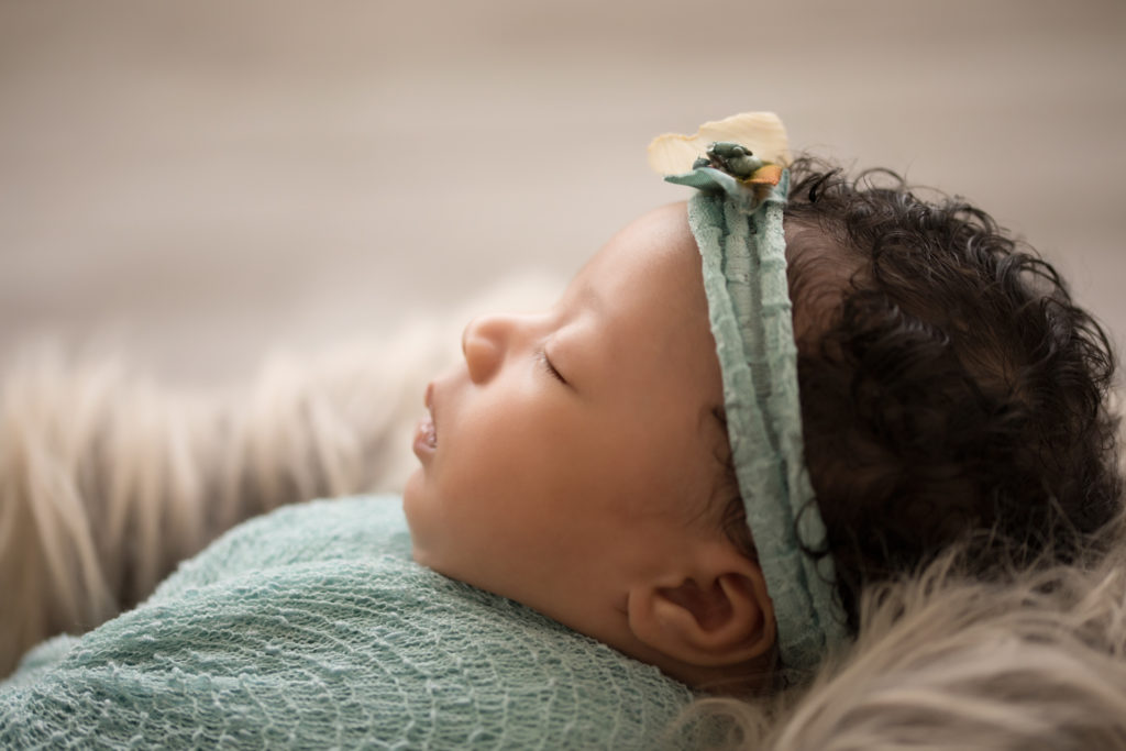 Newborn Girl Ayana close up wrapped in aqua and teal wrap and headband on grey fur with soft light coming from behind her profile face features