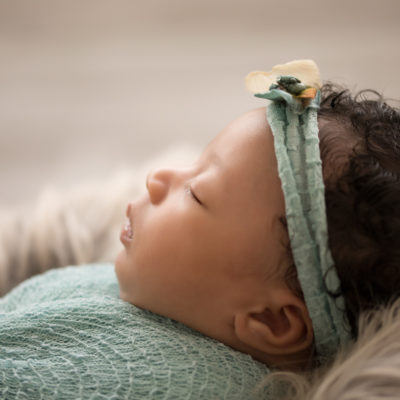 Newborn Girl Ayana close up wrapped in aqua and teal wrap and headband on grey fur with soft light coming from behind her profile face features photos Gainesville FLorida