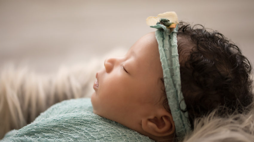 Newborn Girl Ayana close up wrapped in aqua and teal wrap and headband on grey fur with soft light coming from behind her profile face features photos Gainesville FLorida