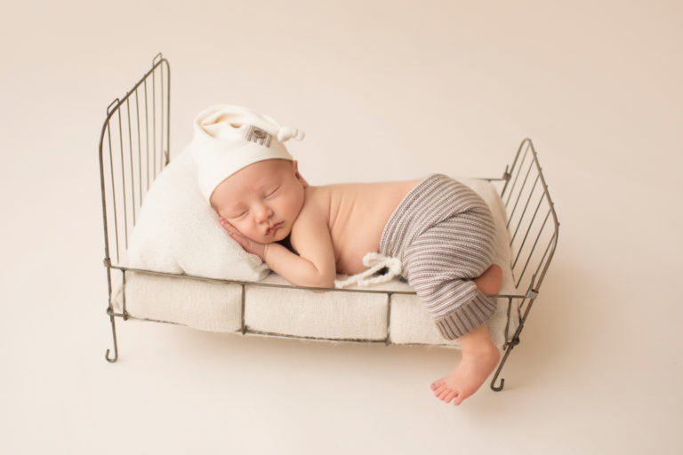 Newborn Baby Jonah with striped knit pants and cream sleepy hat posed on metal bed with leg hanging off newborn photos Gainesville Florida
