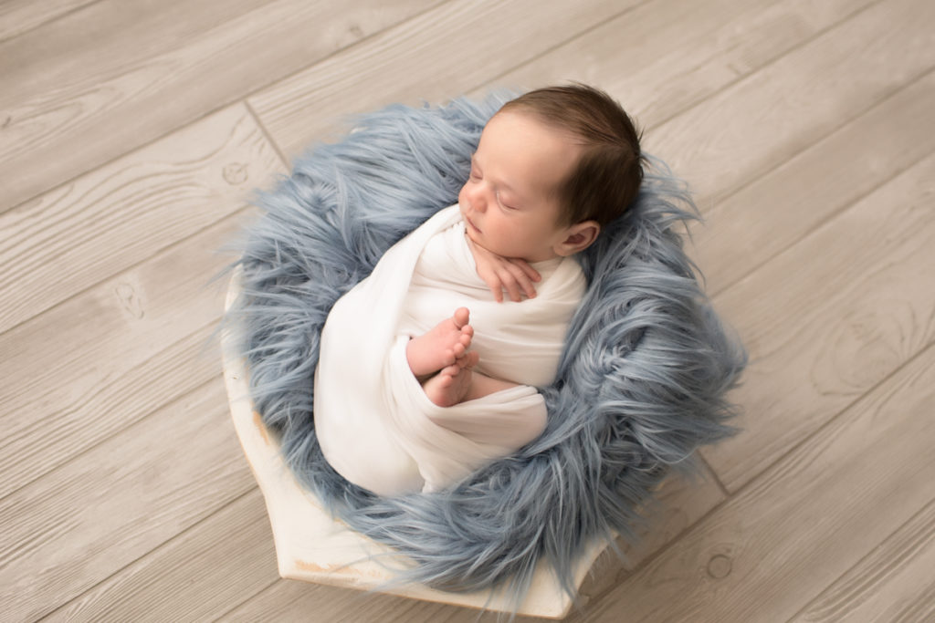 Baby boy Aidan wrapped snuggly with blue fur in white wooden round bowl against wood floor -2