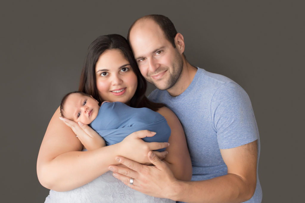 First family portrait mom dad in blues and greys cuddling newborn baby with eyes open to camera