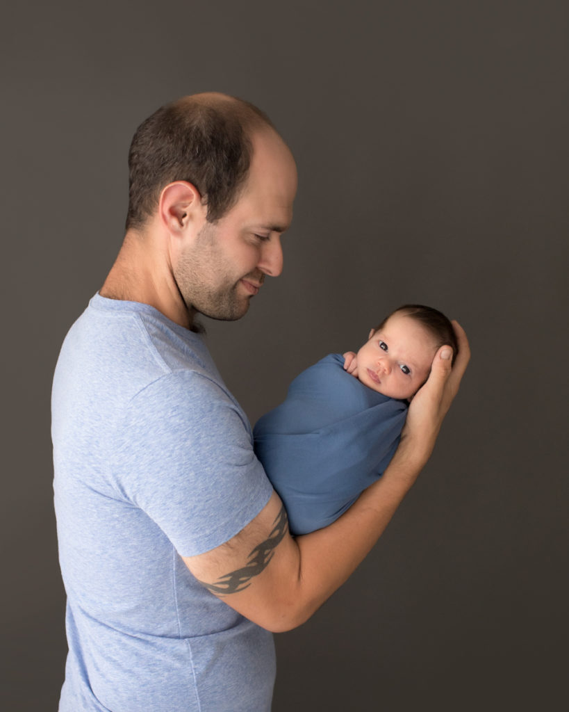 Profile dad with tatoo admiring his newborn baby boy with eyes open to camera