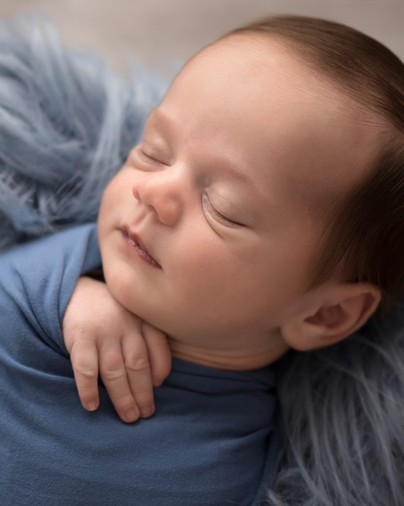 Newborn boy Aidan face close up and swaddled snuggly in blue wrap against blue fur