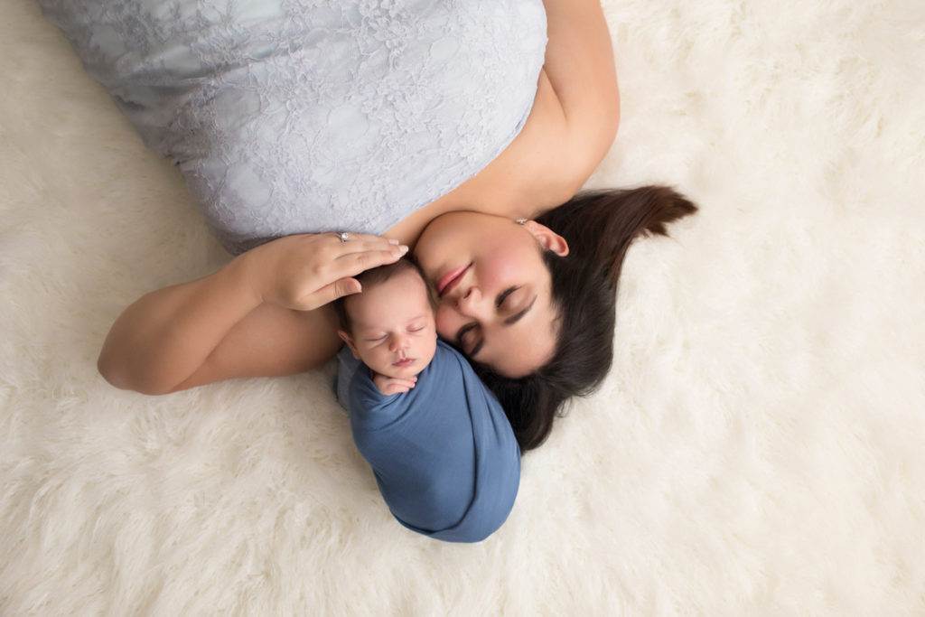 Beautiful new Mom in grey lace cuddling newborn Aidan wrapped in blue on white fur rug