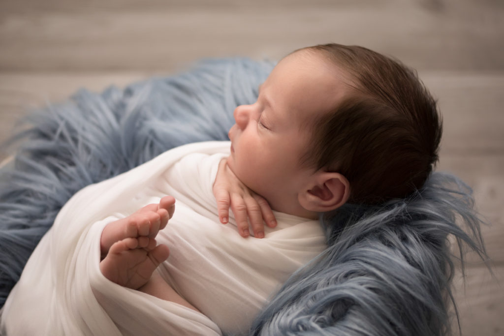 Baby boy Aidan face profile and wrapped snuggly with blue fur in white wooden bowl against wood floor