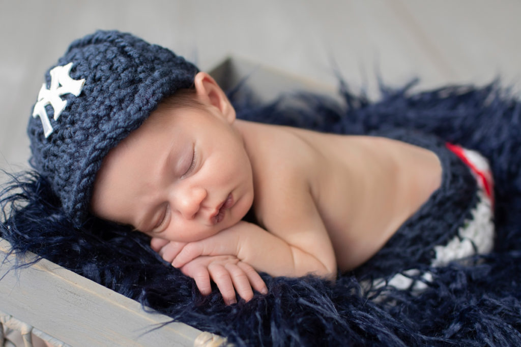 Newborn boy Aidan in baseball pants outfit and cap sleeping on blue fur iin a wooden box prop