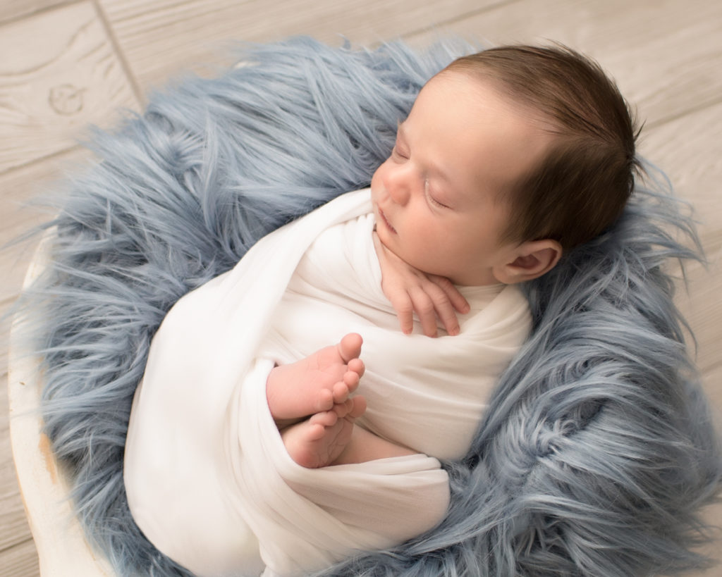 Baby boy Aidan wrapped snuggly with blue fur in white wooden bowl against grey wood floor