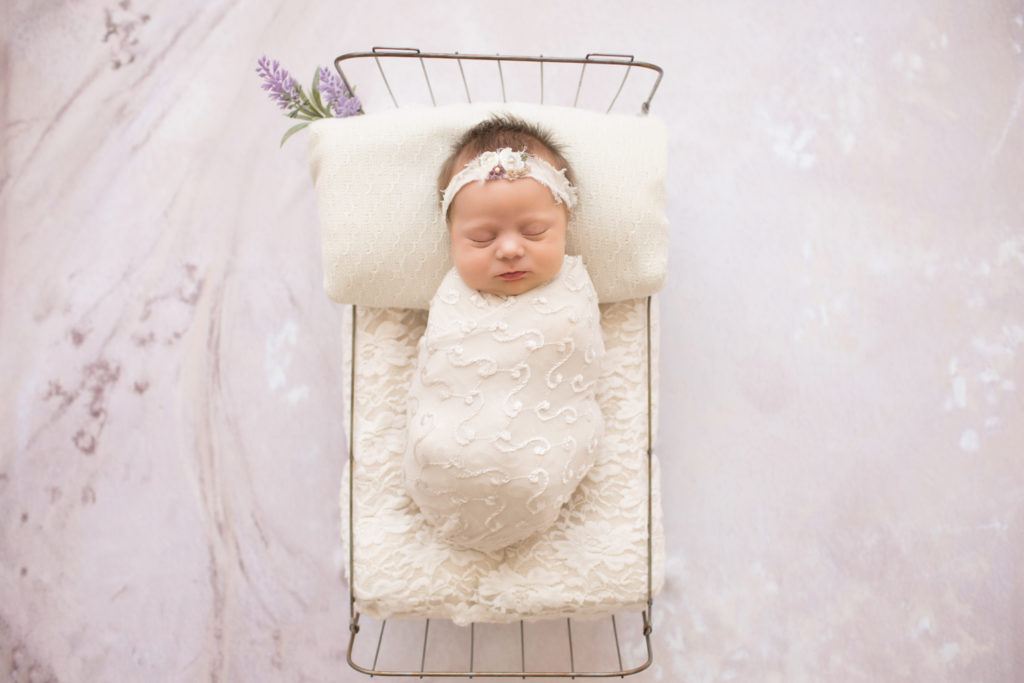 Newborn girl wrapped in cream lacey knit posed in baby bed sleeping against lilac flower background