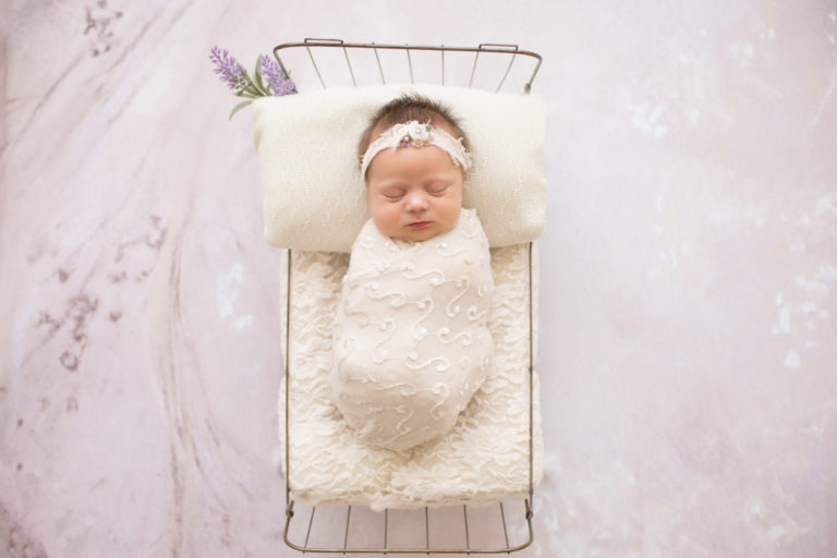 Newborn girl wrapped in cream lacey knit posed in baby bed sleeping against lilac flower background photos Gainesville FLorida
