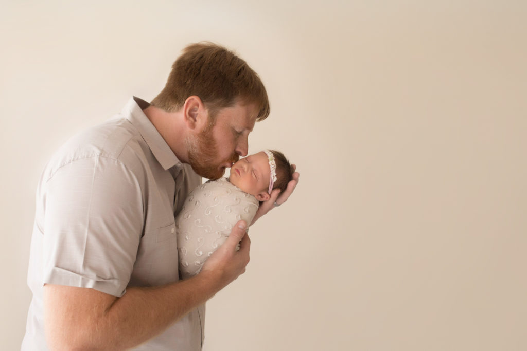 Dad in beige cuddling and kissing newborn girl wrapped in cream in football hold