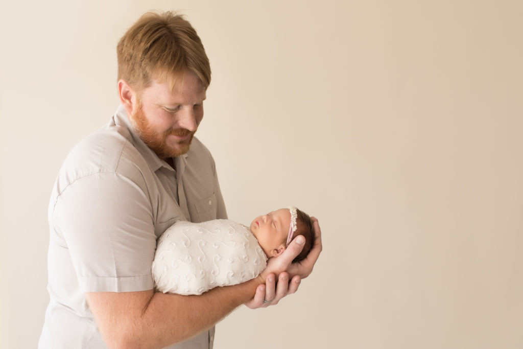 Dad in beige cuddling and admiring newborn girl wrapped in cream in football hold