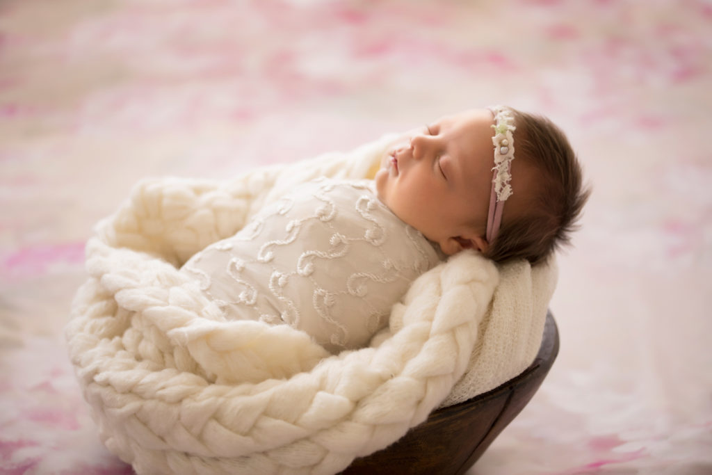 Newborn girl wrapped in pretty cream knit fabric sleeping in brown wooden bowl with chunky knit blanket