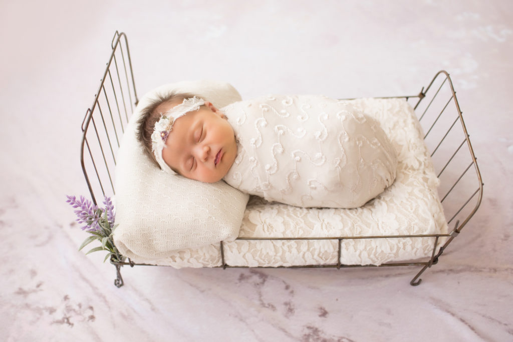 Newborn girl wrapped in pretty cream knit fabric asleep and posed on baby bed with lace and purple flowers