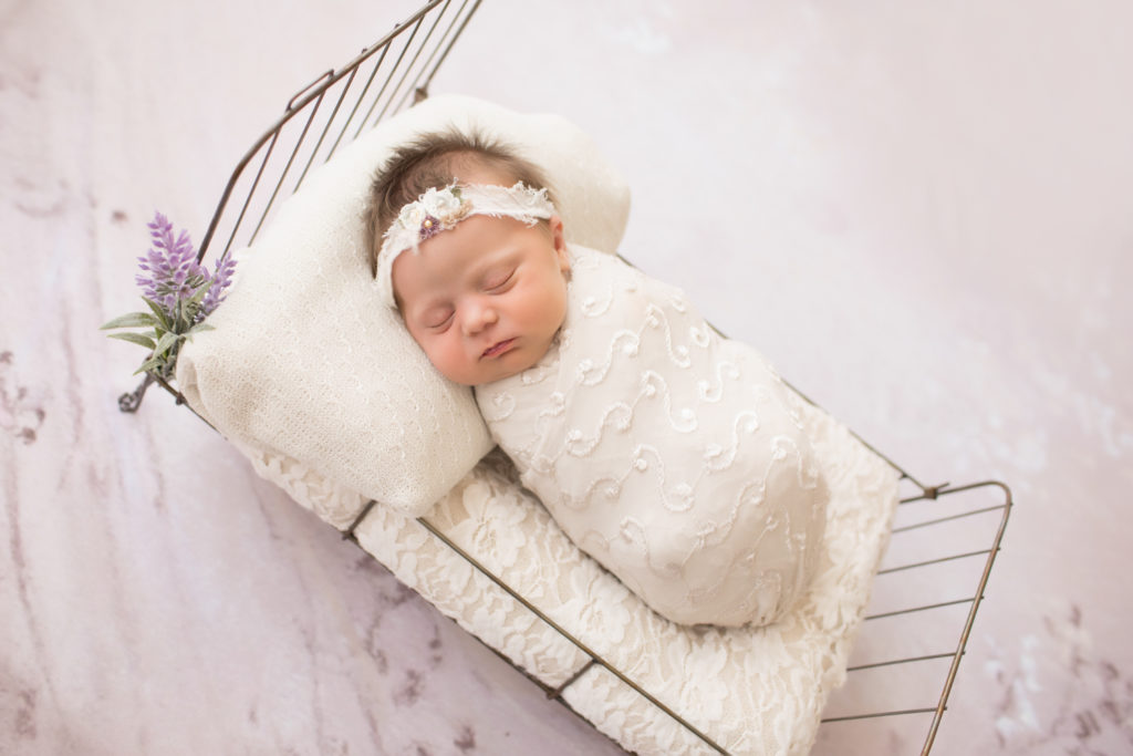 Newborn girl wrapped in pretty cream knit fabric sleeping on baby bed with lace and purple flowers