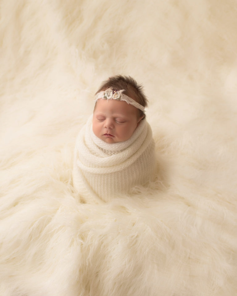 Newborn baby girl wrapped in white knit standing upright like potato sack against white fur photos Gainesville FLorida
