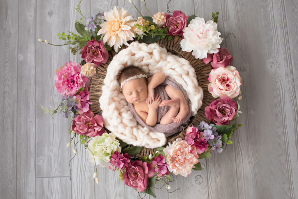 Newborn baby girl wrapped in pink in basket of rosey flowers with pearl headband against grey floor
