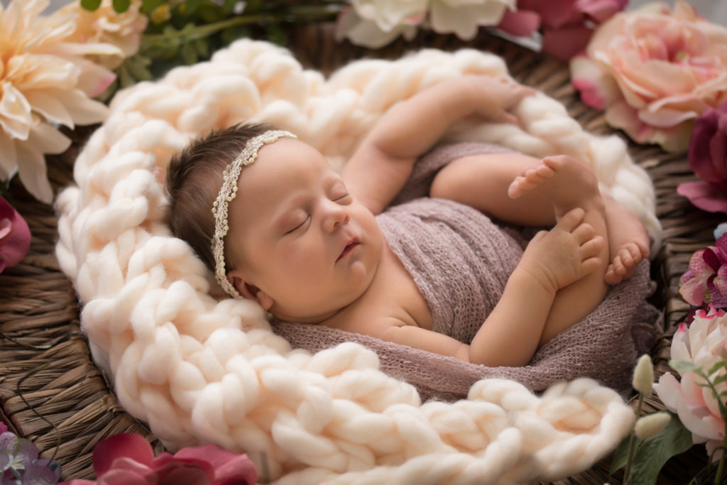 Newborn baby girl wrapped in pink in basket of rosey flowers with pearl headband on head