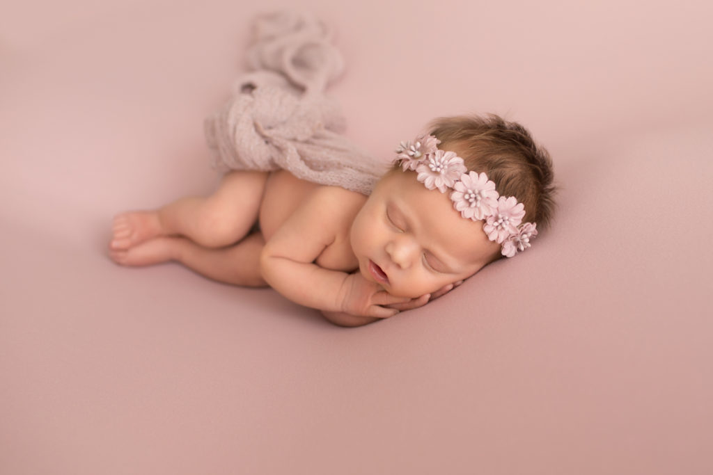 Newborn naked baby girl wrapped in pink snuggling on blush blanket with floral crown on head
