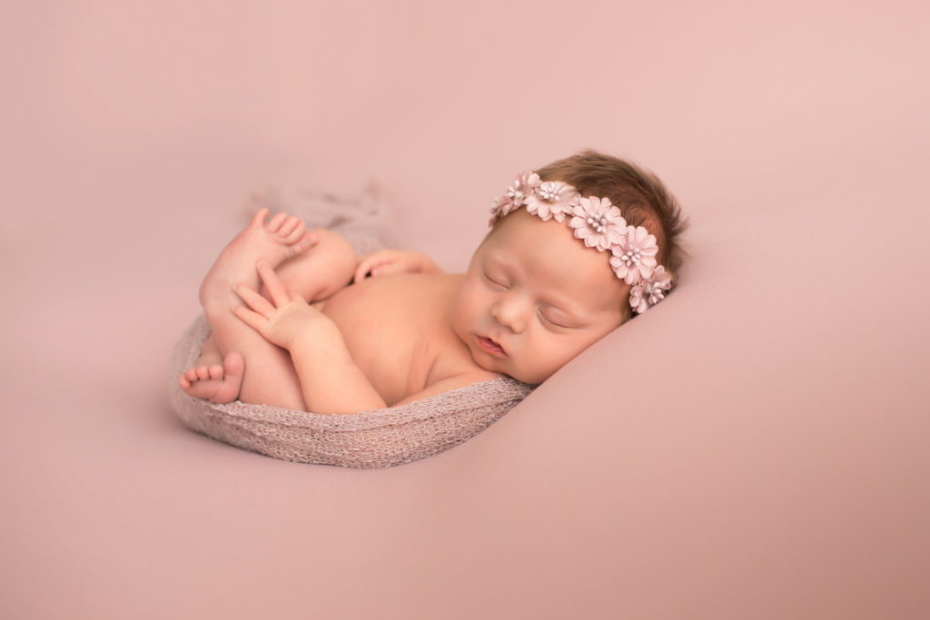 Newborn naked baby girl wrapped in pink on blush blanket with floral crown on head