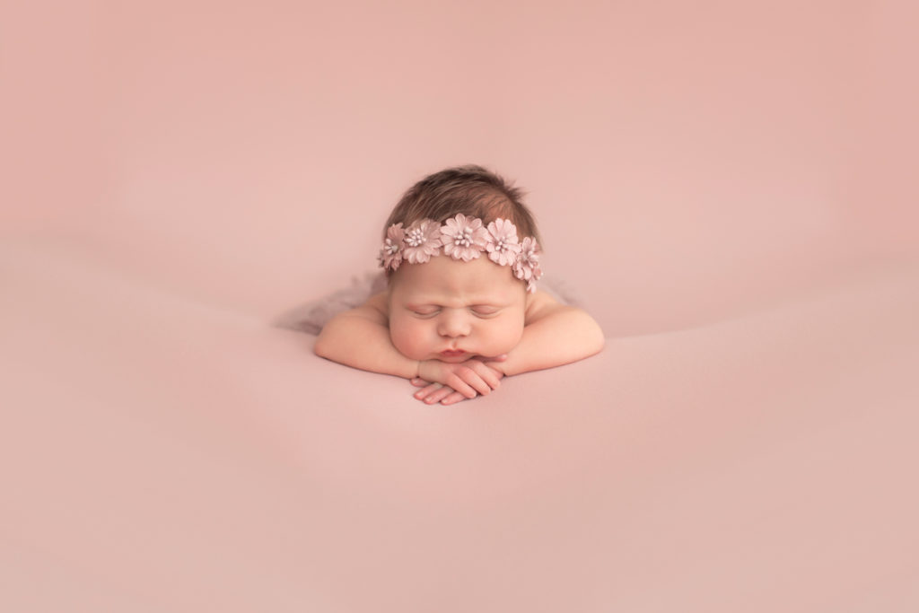 Newborn baby girl with chubby cheeks posed and sleeping on pink blanket with chin resting on her wrists flower crown on head