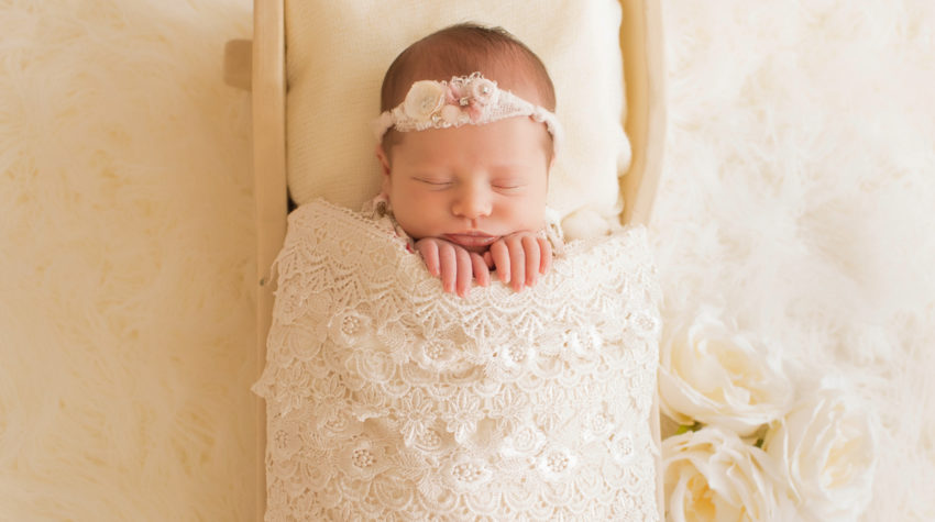 Newborn baby girl Bailey lying in cream bed wrapped in ivory lace with baby fingers wrapped on lace blanket and cream roses Gainesville FL Andrea Sollenberger Photography