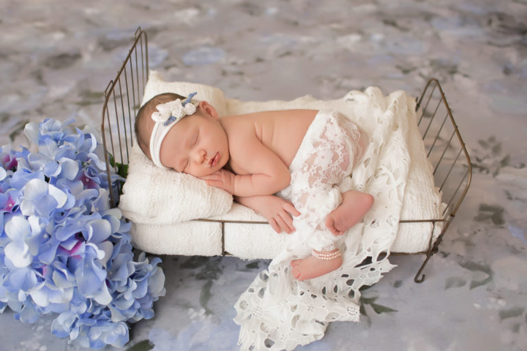 Newborn baby girl Bailey lying on side on metal bed white blanket white lace blue flowers pearls and floral white headtie Gainesville FL Andrea Sollenberger Photography