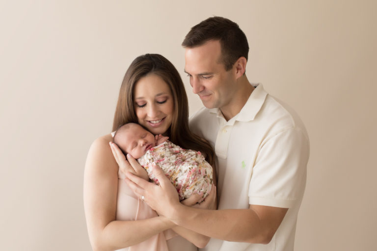 Gorgeous mom handsome dad neutral color portrait smiling looking down and cuddling newborn baby girl Bailey in pink floral wrap Gainesville FL Andrea Sollenberger Photography
