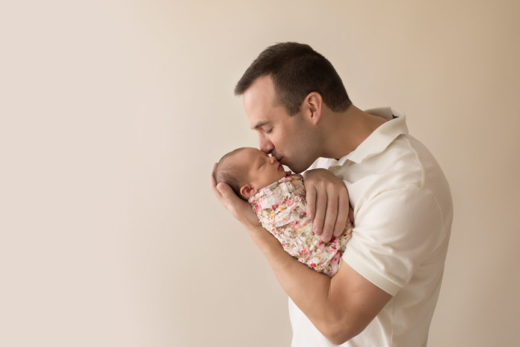 Handsome dad neutral color portrait kissing and cuddling newborn baby girl Bailey in pink floral wrap Gainesville FL Andrea Sollenberger Photography