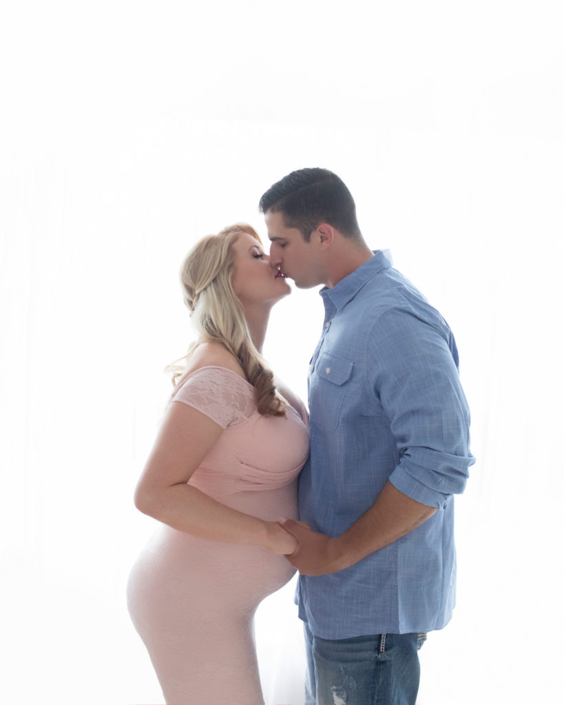 Maternity photos Christina mom face to face with her man Jamie kissing against backlight profile beautiful belly bump wearing mermaid style full length pale pink lace gown Gainesville Florida