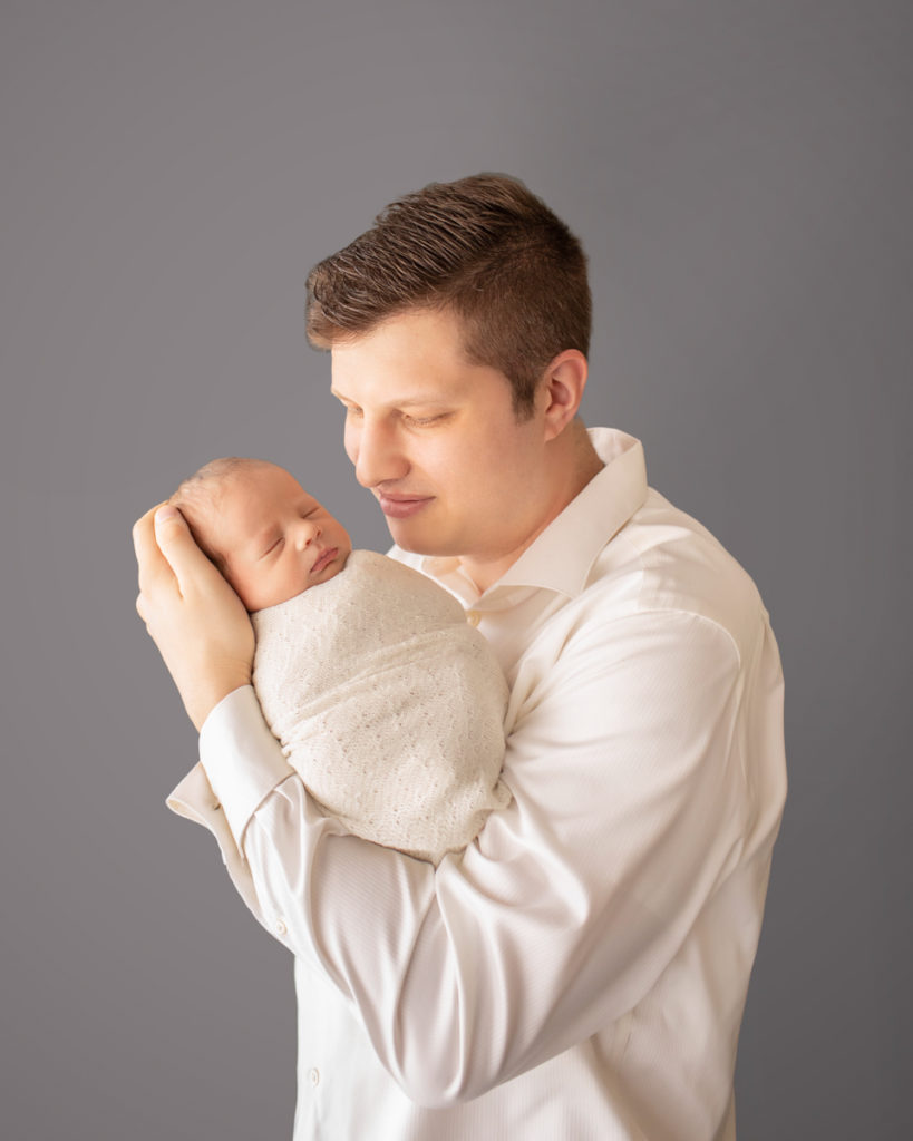 Dad looking gazing and cuddling newborn baby Rowan dressed in white against grey Gainesvile Florida Photos