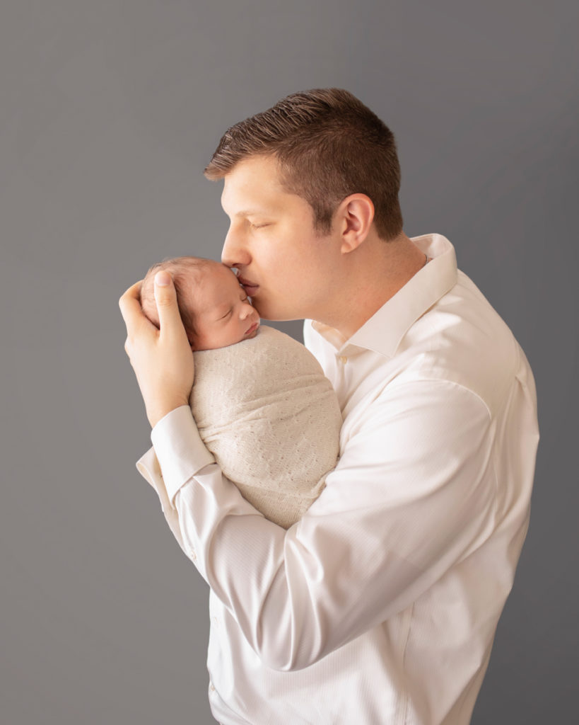 Dad looking kissing and cuddling newborn baby Rowan dressed in white against grey Gainesvile Florida Photos