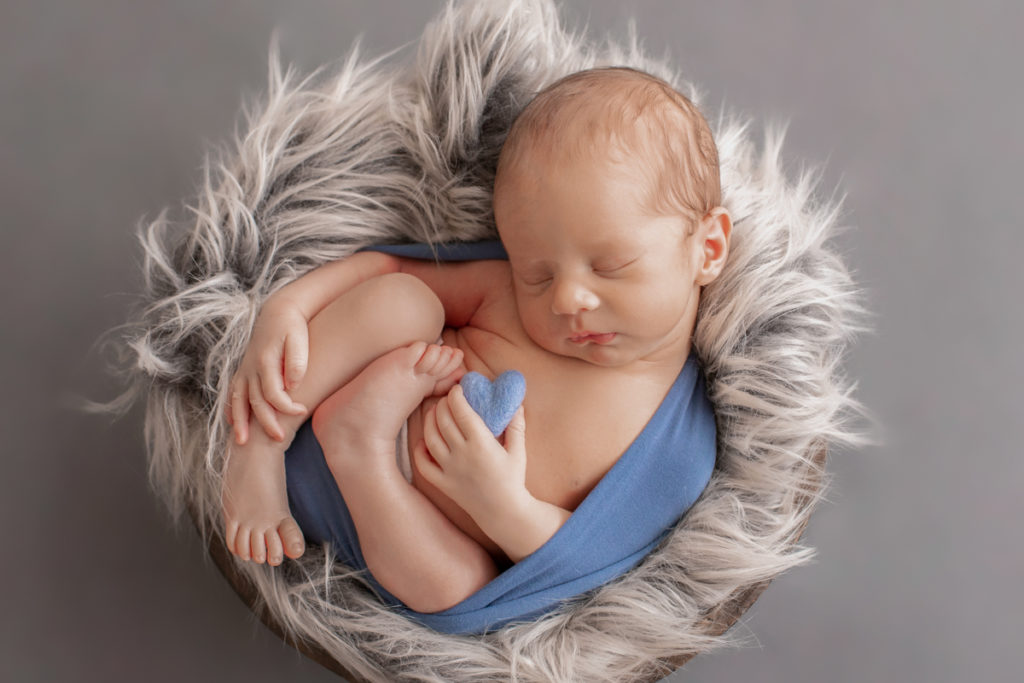 Newborn Baby Rowan wrapped in blue holding tiny heart in hand posed in brown bowl with grey fur Gainesvile Florida Photos