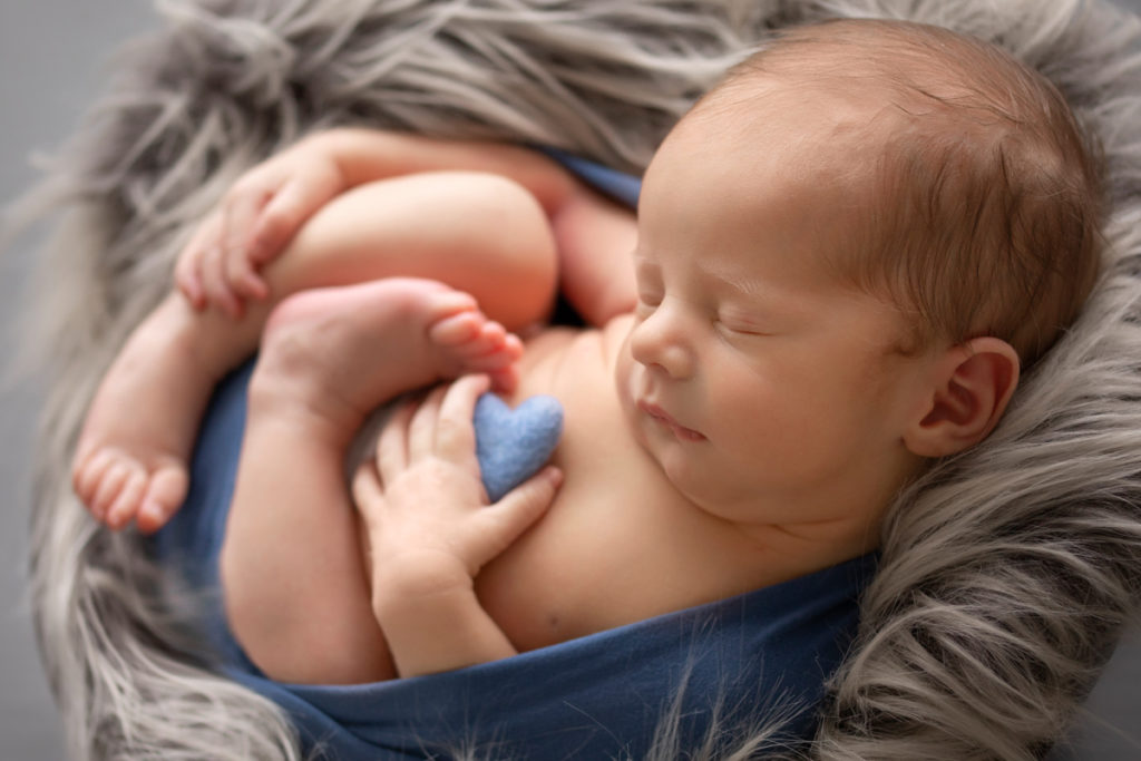 Newborn Baby Rowan wrapped in blue holding tiny blue felt heart in hand posed with grey fur side view Gainesvile Florida Photos